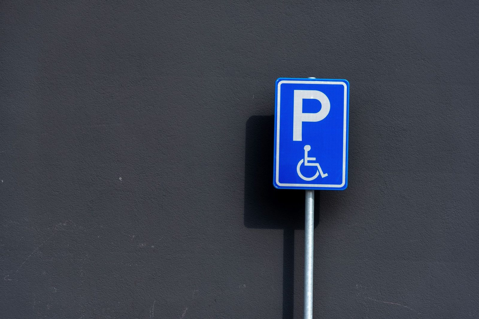 Señal azul de estacionamiento para personas con discapacidad montada en un poste metálico frente a una pared oscura.