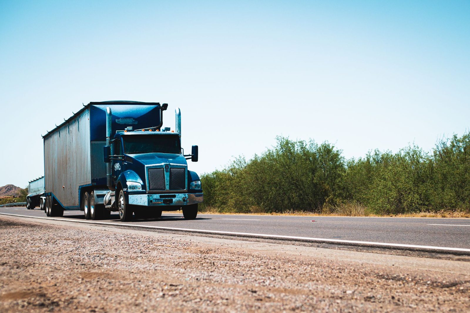 A blue semi truck traveling down a rural highway under a clear blue sky.