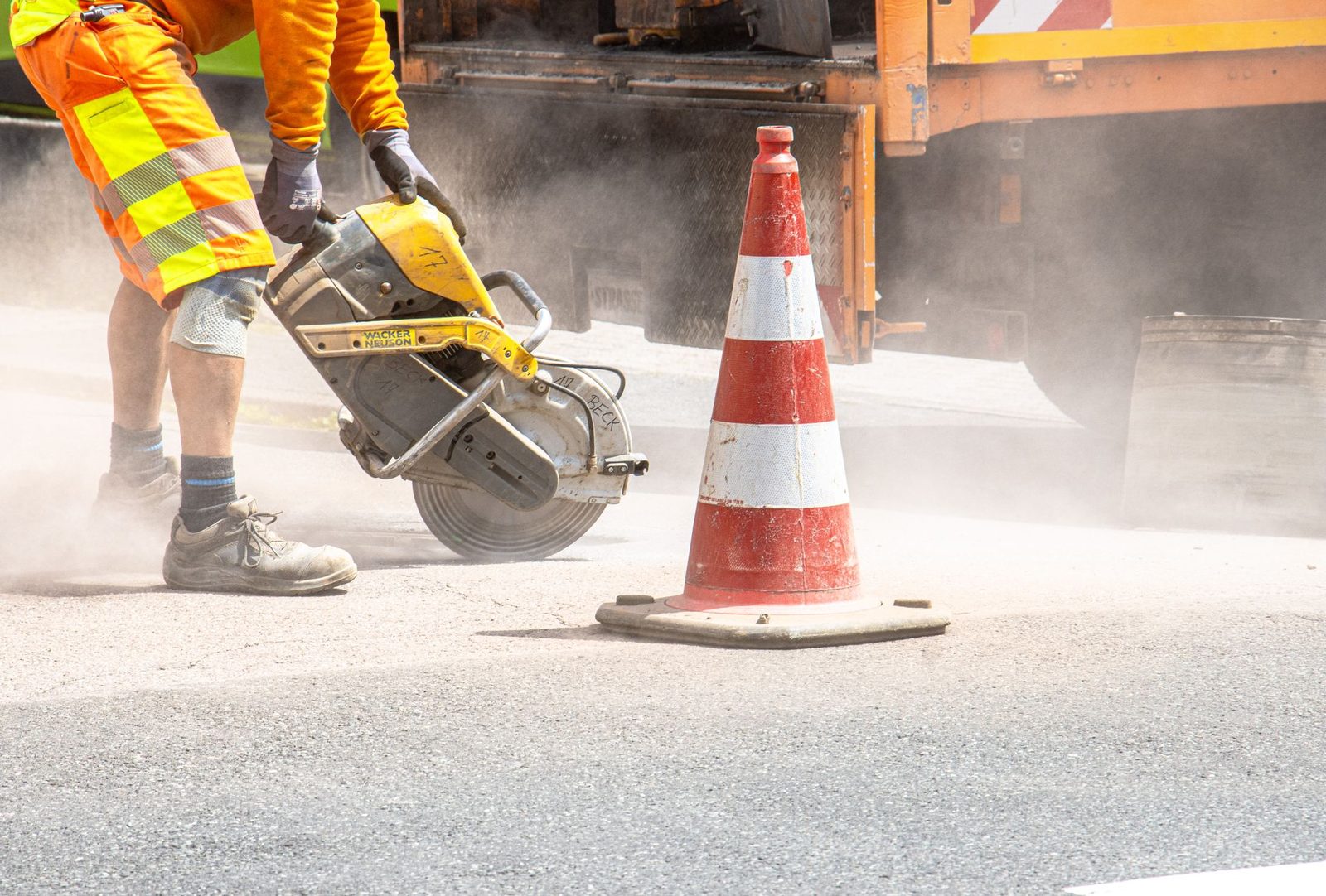A construction worker cuts asphalt with a large saw beside a traffic cone on a street.