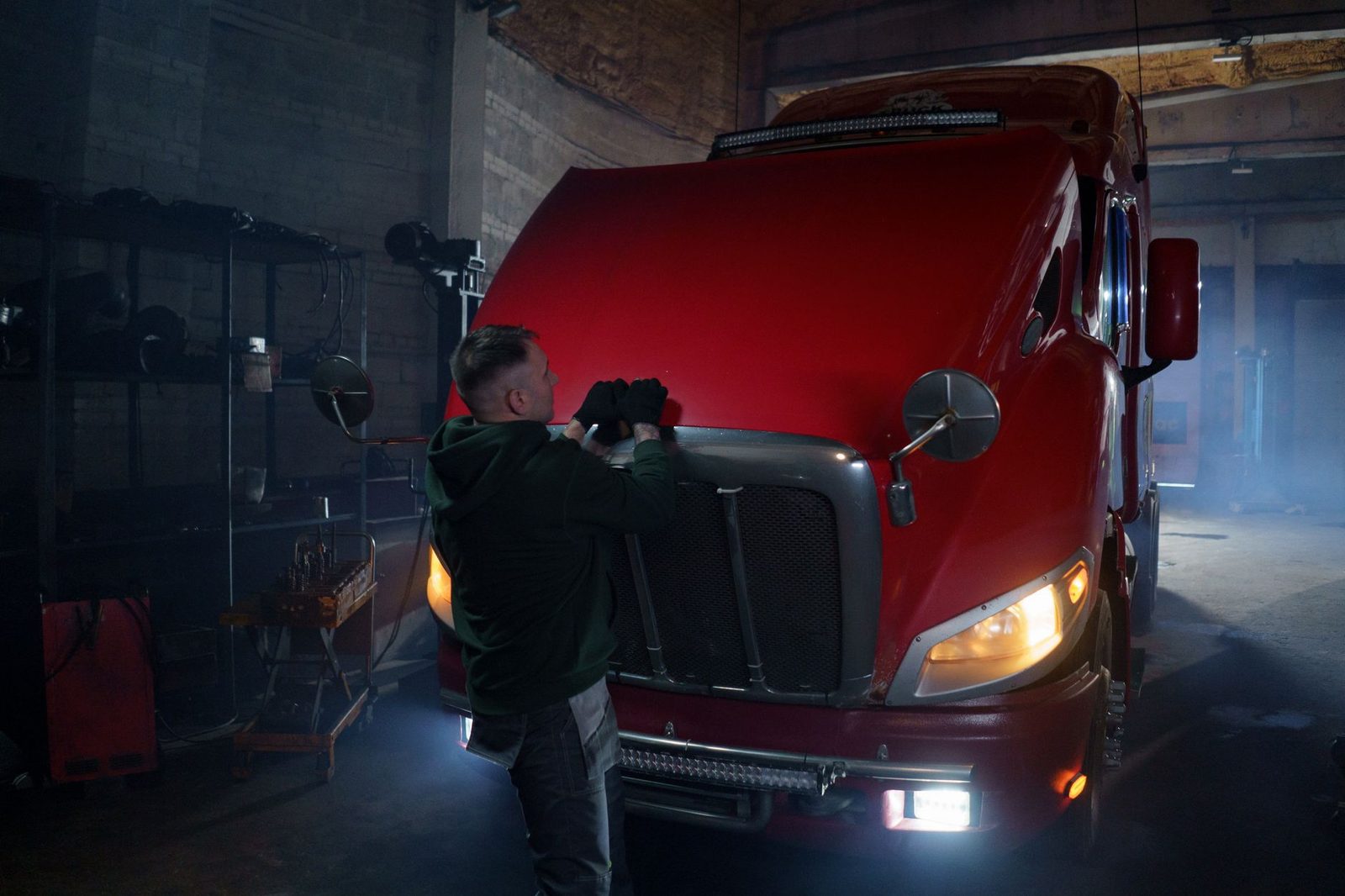 A mechanic in gloves works under the hood of a red truck in a dimly lit garage.