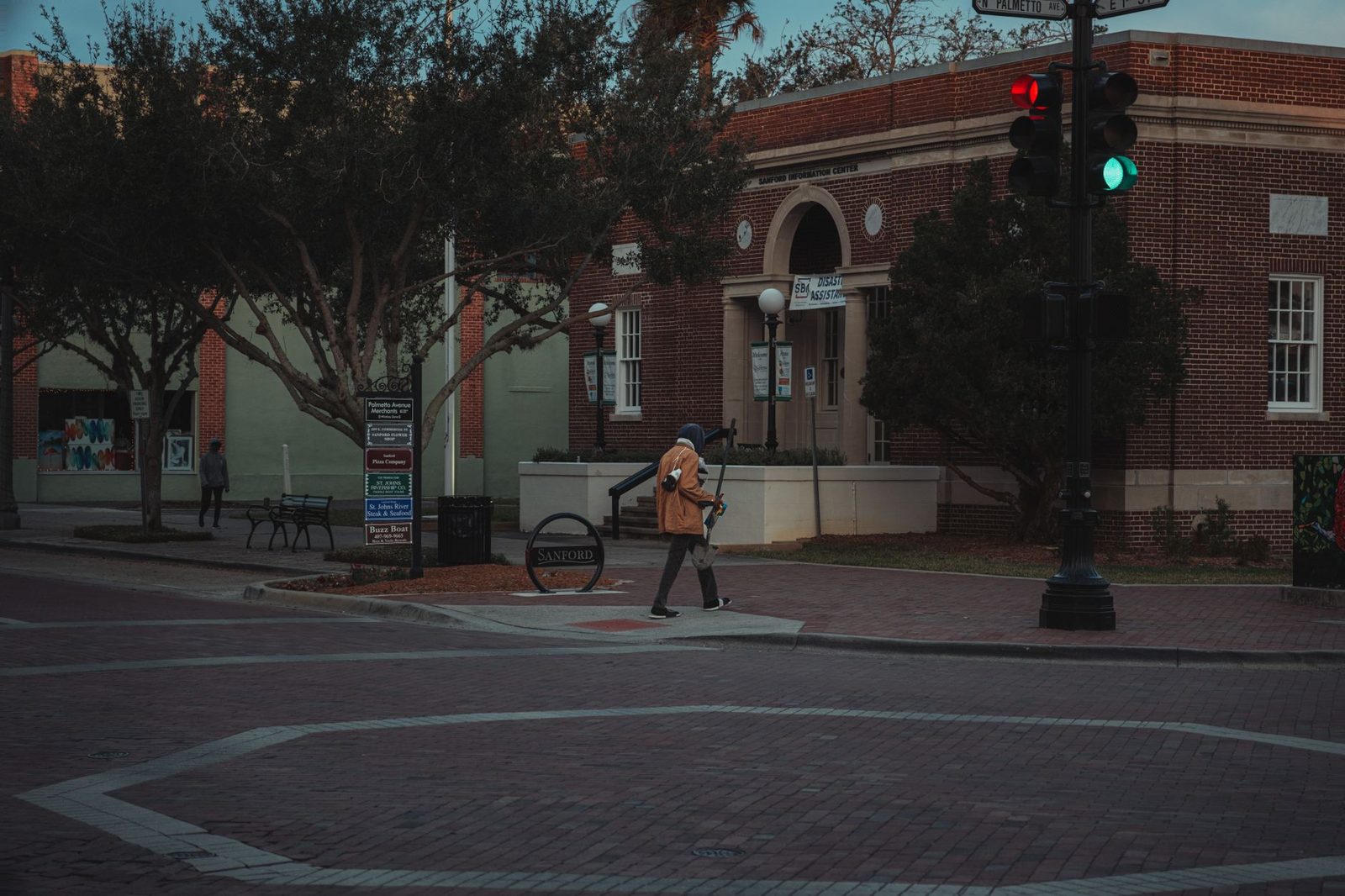 A pedestrian walks through a city street intersection at twilight, past brick buildings and trees.