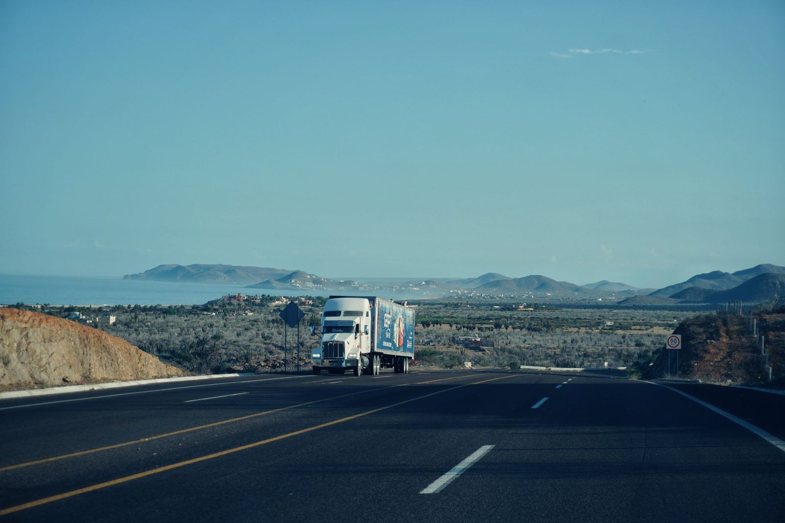 A semi-truck drives along a scenic highway with mountains rising in the background under a clear sky.