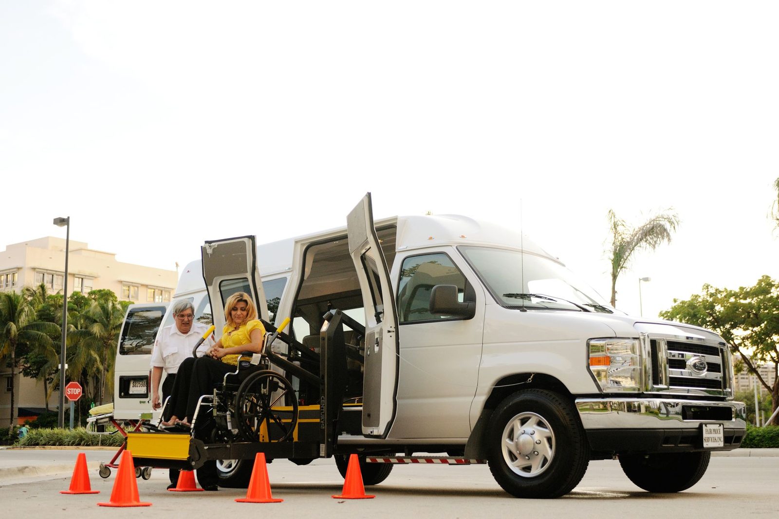 A woman in a wheelchair boards an accessibility van using a lift while a driver assists her.