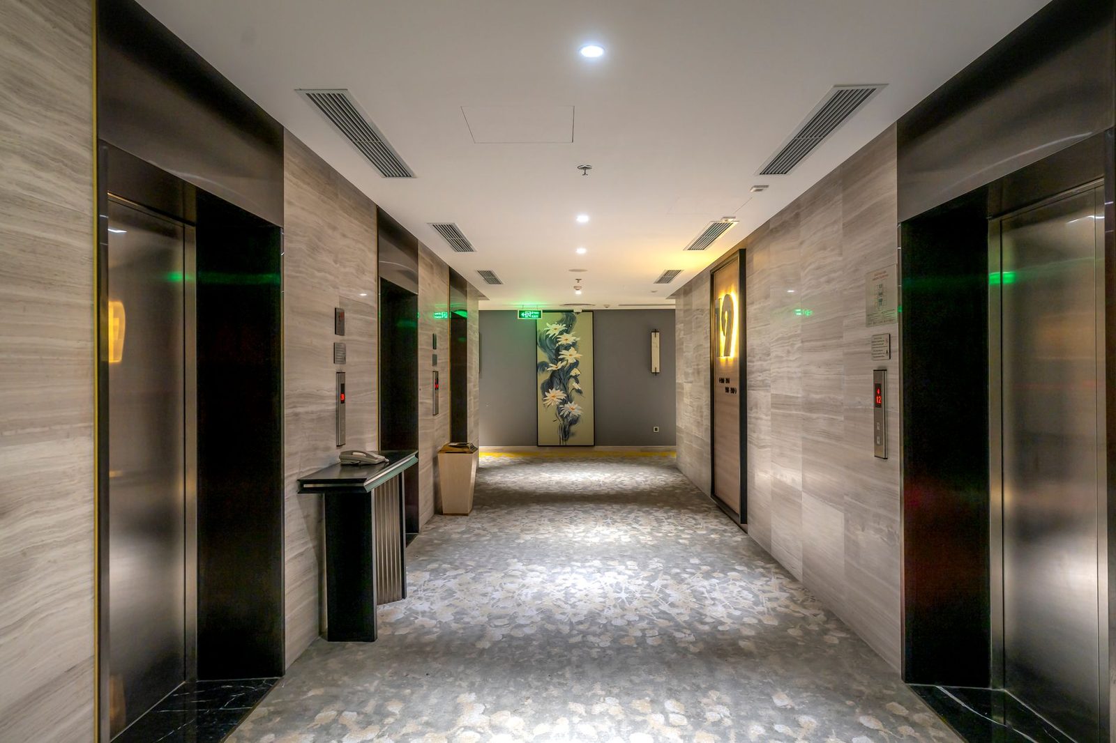 Empty hotel hallway with elevators and modern decor, lit by warm overhead lights.