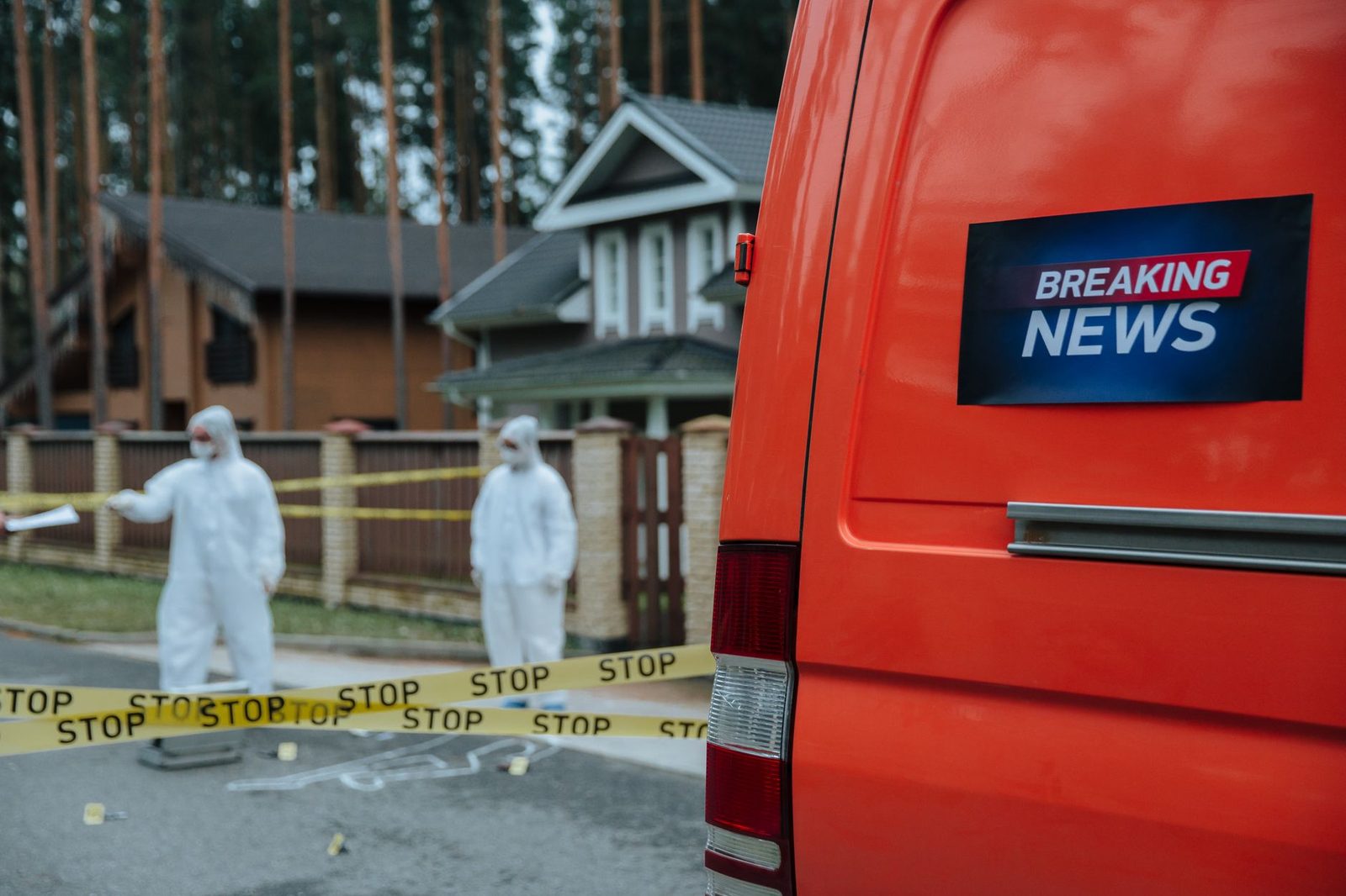 Forensic investigators in protective suits work at a marked-off scene beside concrete houses.