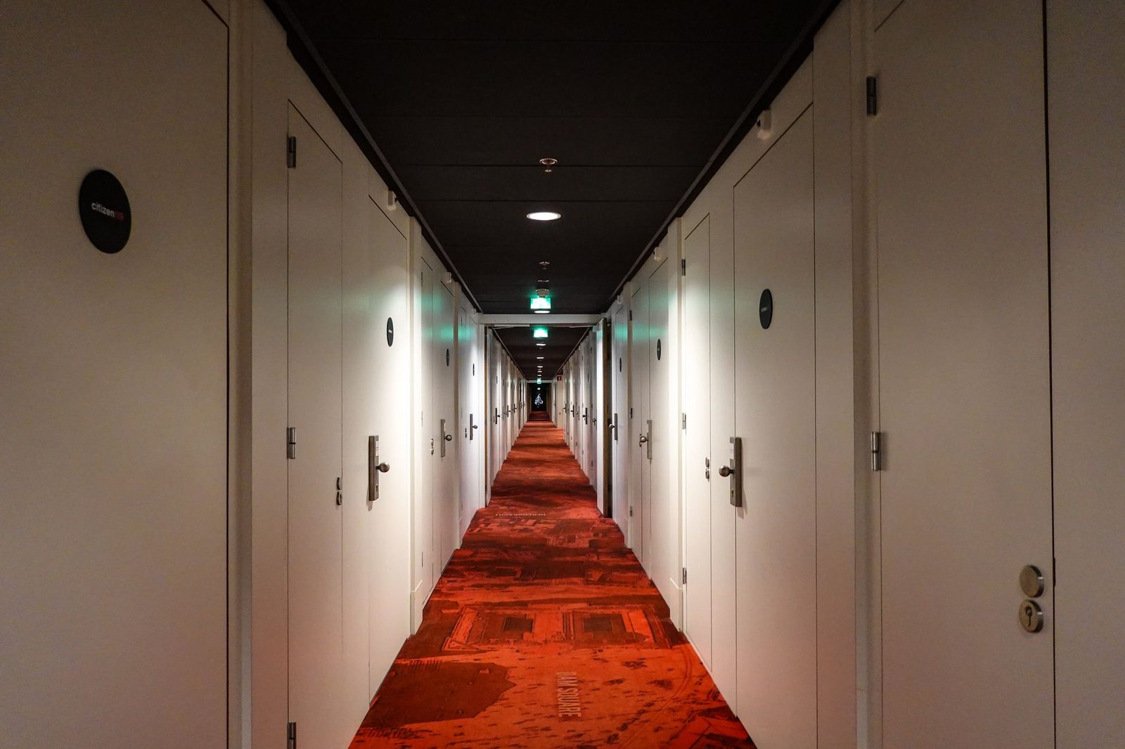 Long, symmetrical hotel hallway with red carpet, closed white doors, and warm lighting.