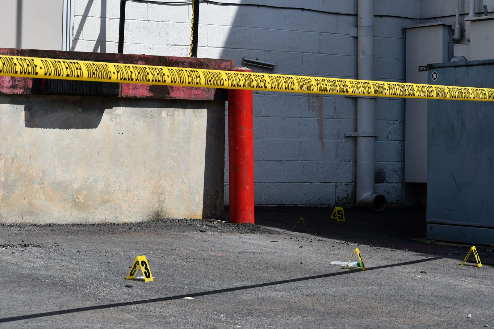 Escena callejera iluminada por el sol, con cinta amarilla de precaución y marcadores numerados de evidencia en una escena del crimen.